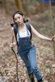 A woman in overalls and a backpack standing in the woods.